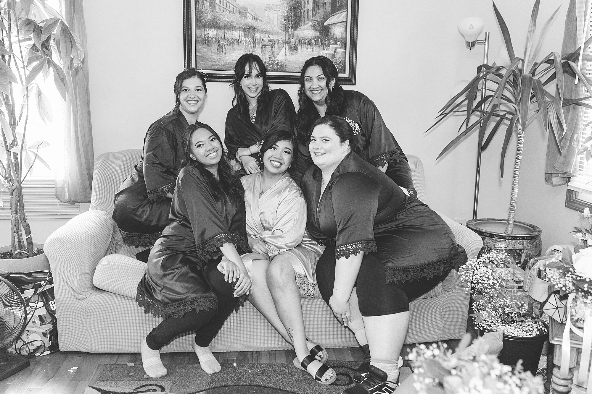 bridesmaids and bride are sitting on a coach taking a group photo in a living room 