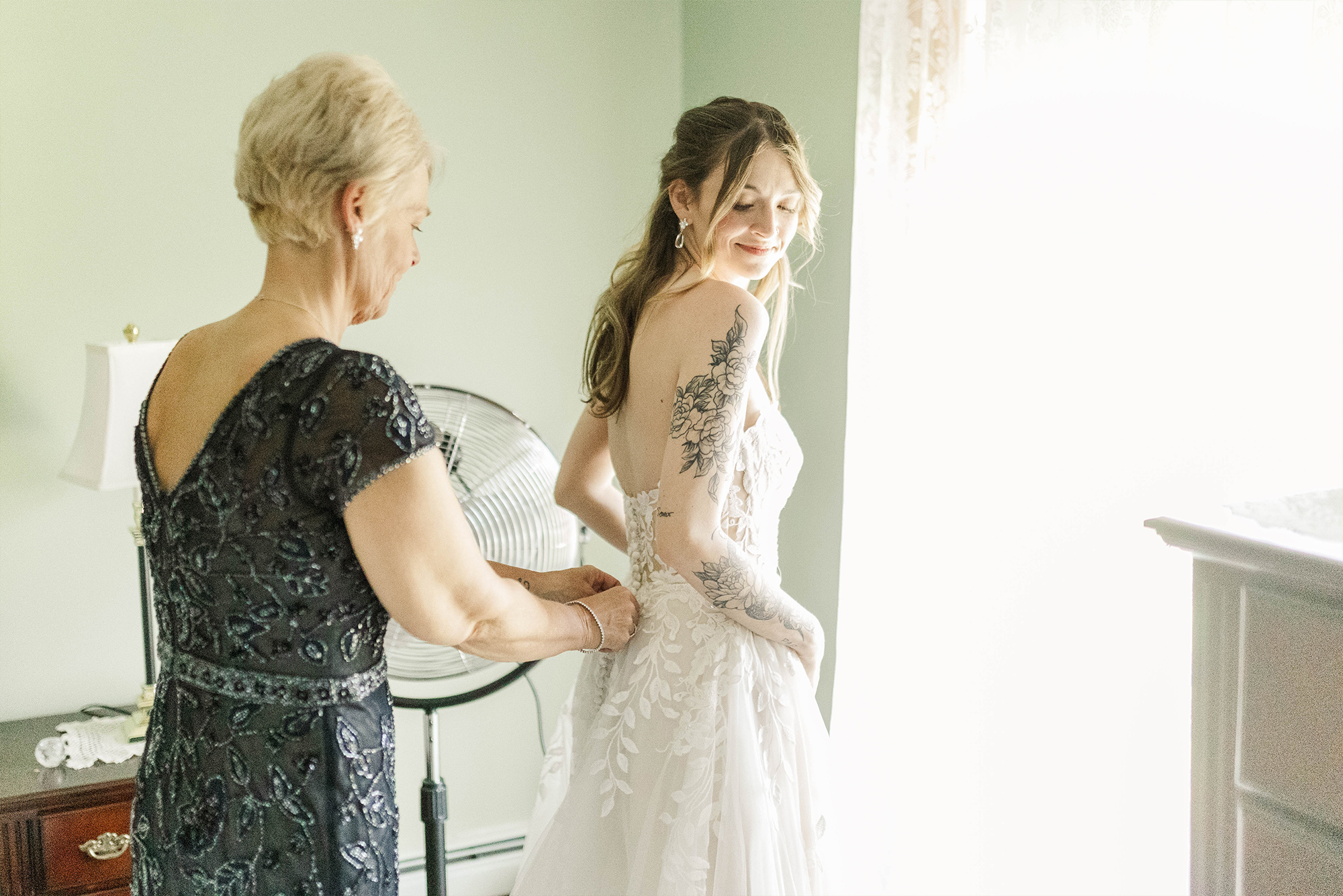 mother helping bridge get into wedding dress next to window