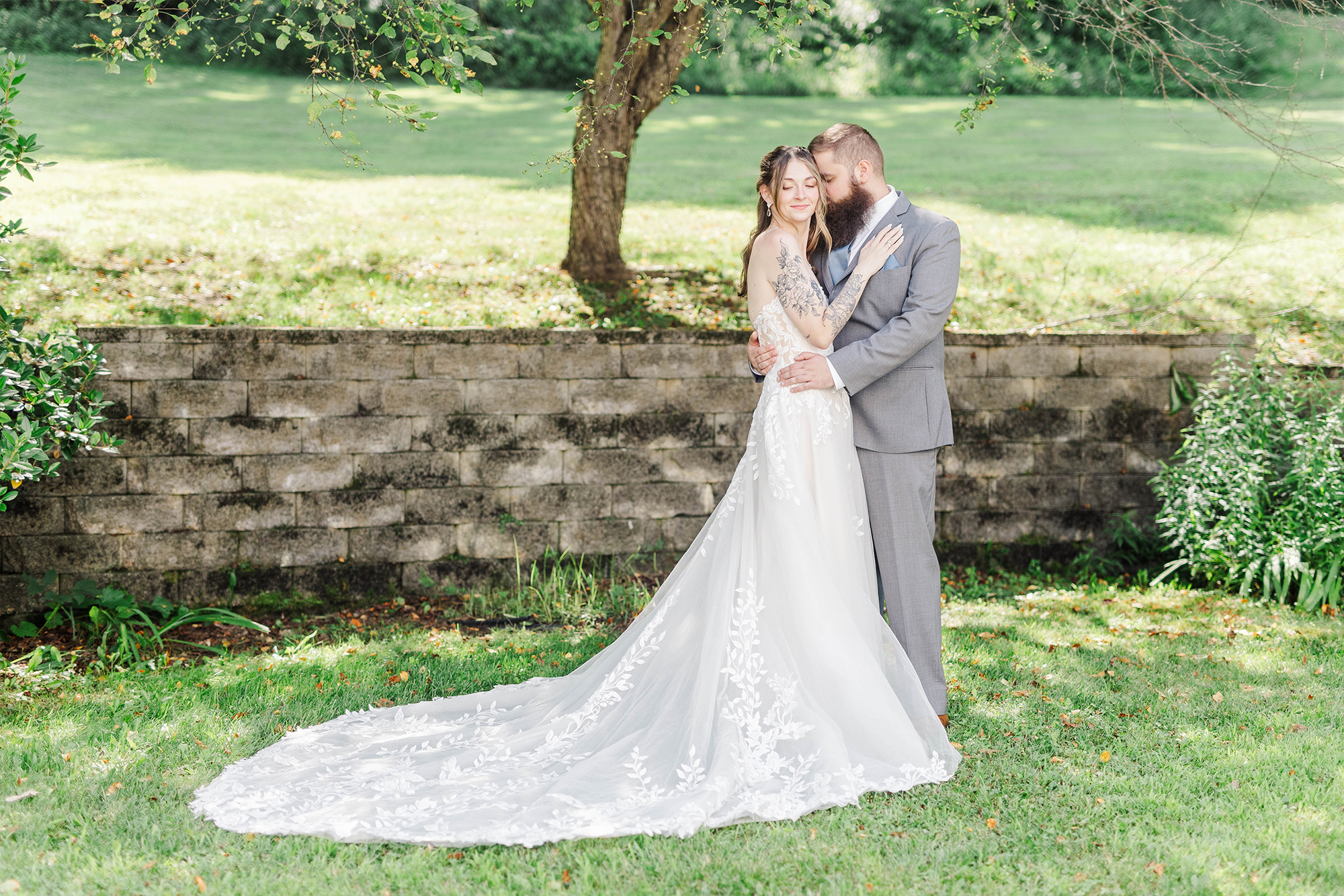 bride and groom embracing while standing under tree with large dress train