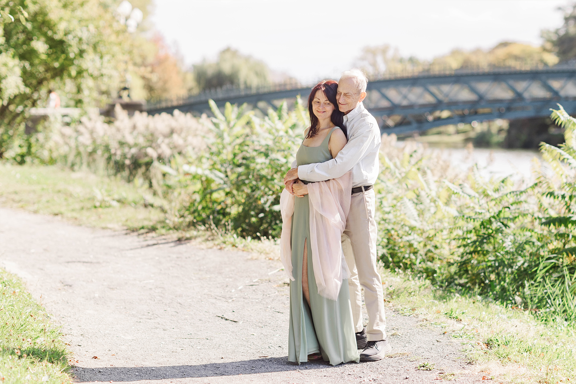 couple holding each other on path with tall greenery and bridge