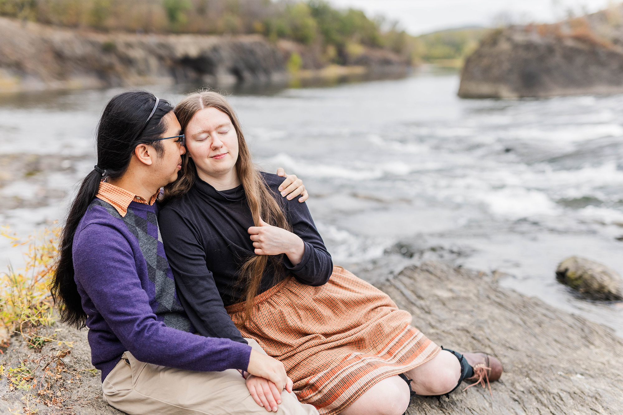 couple sits on rocks holding each other by water