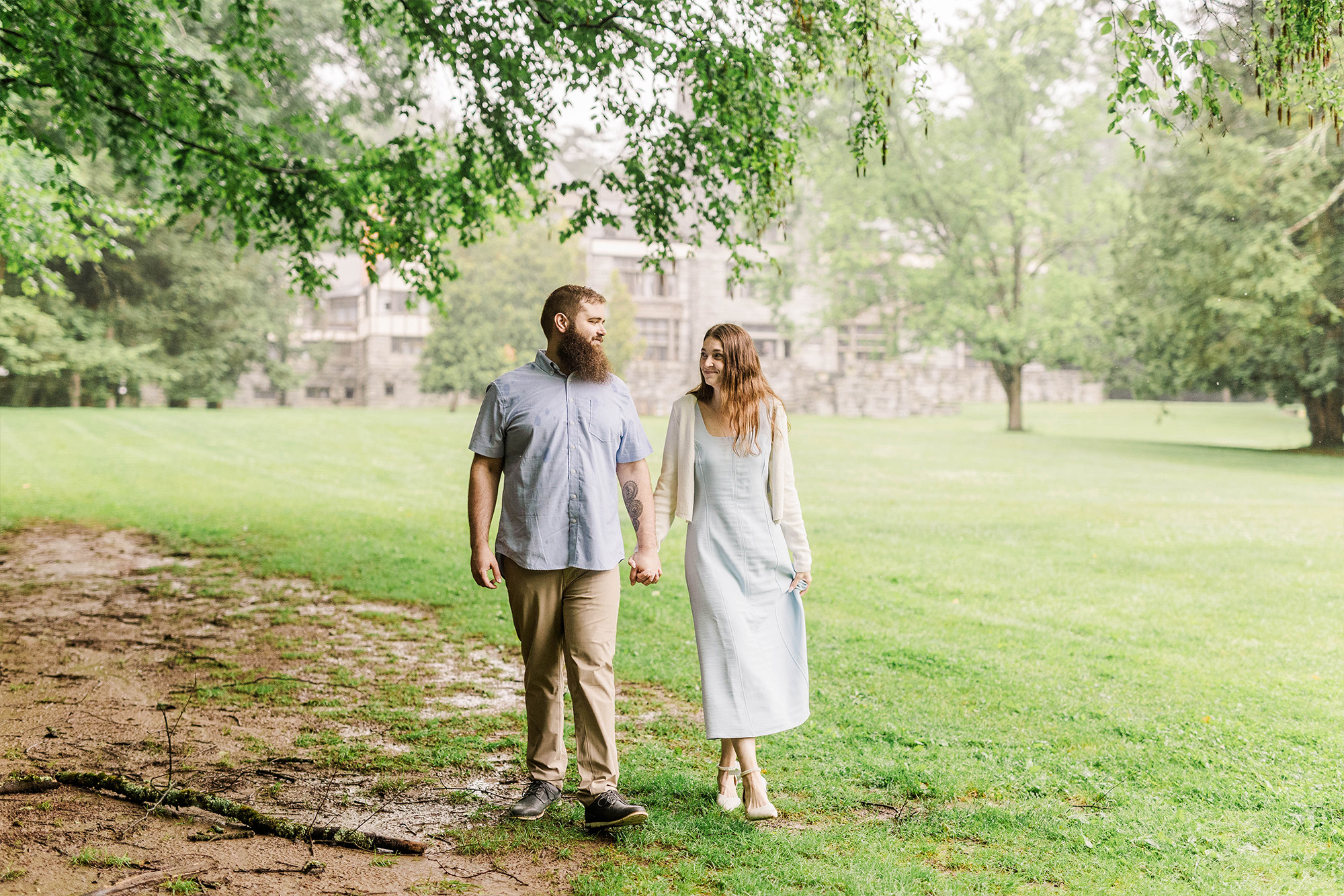 couple walking hand in hand underneath tree with mansion in background