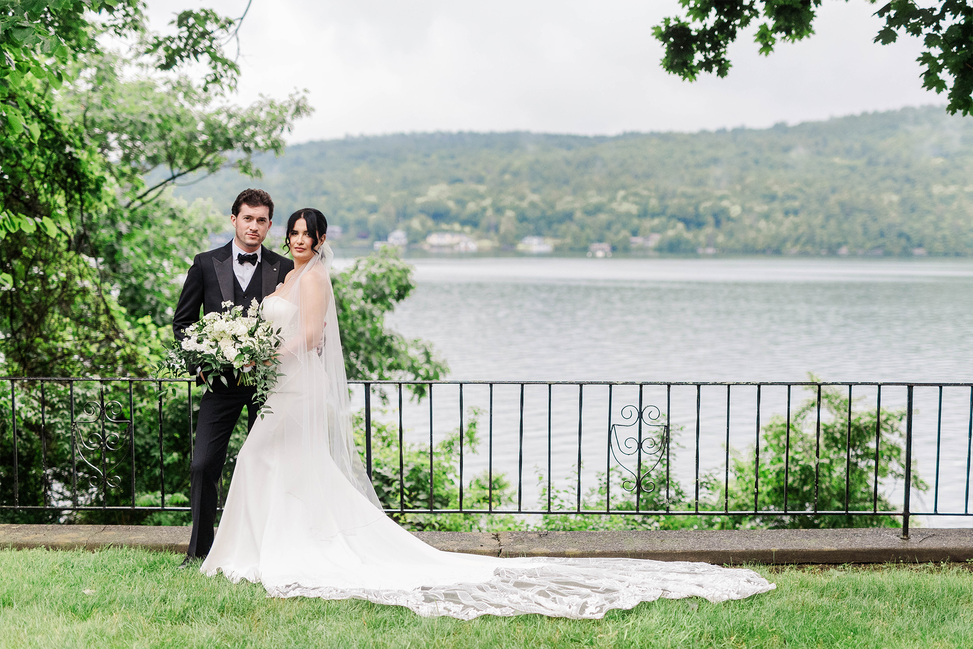 bride and groom standing in front of railing with bridal bouquet with lake backdrop and trees