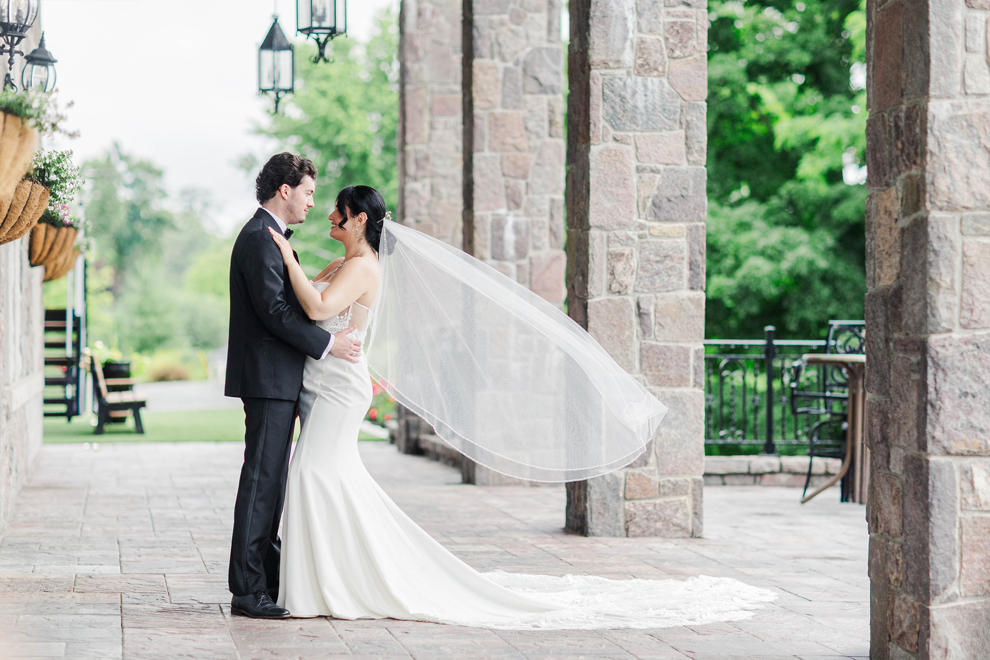 bride and groom holding each other on outdoor patio with stone pillars, hanging lanterns and greenery