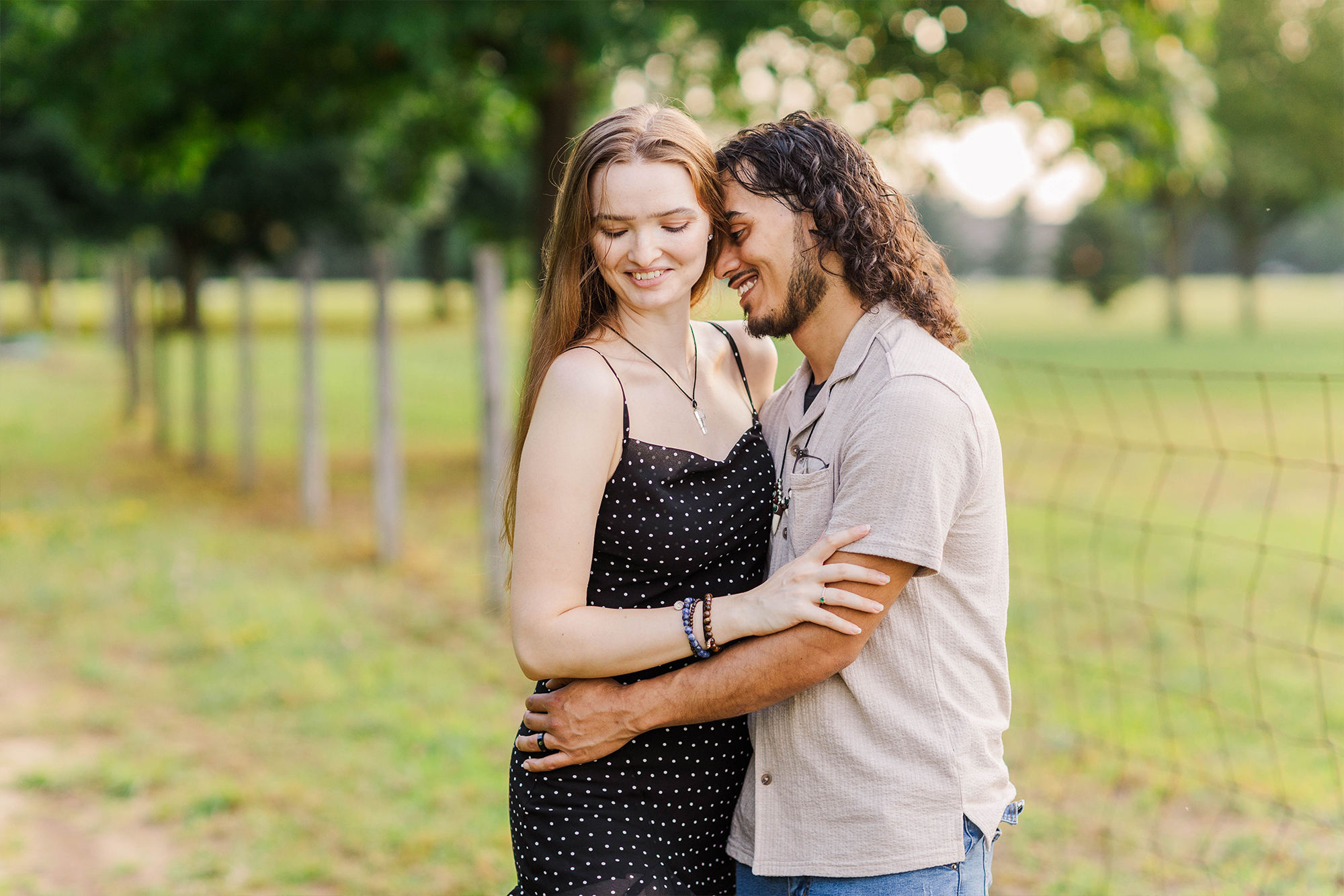 boyfriend and girlfriend holding each other next to fence with trees and golden hour glow