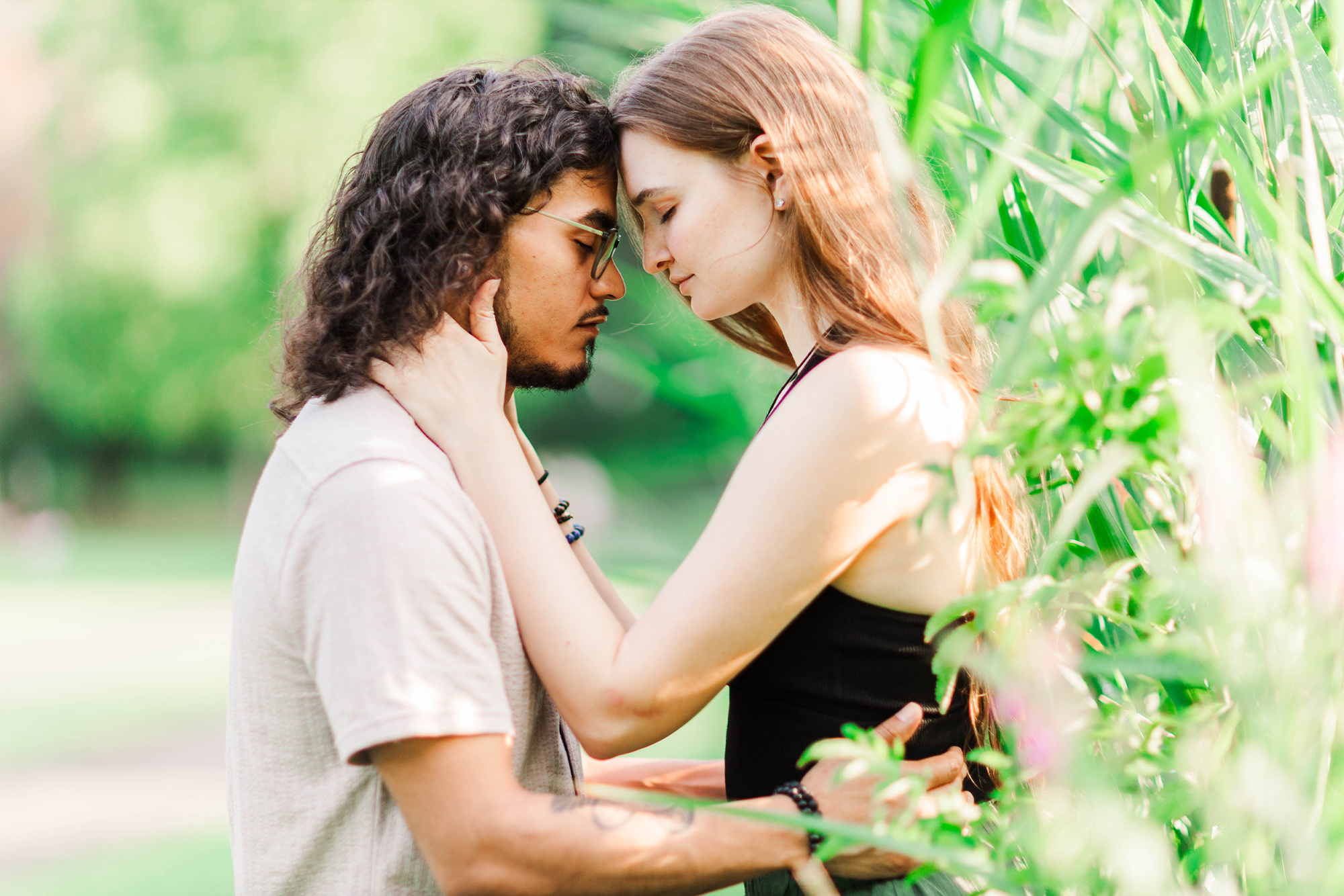 boyfriend and girlfriend with foreheads pressed against each other standing near tall grass in park