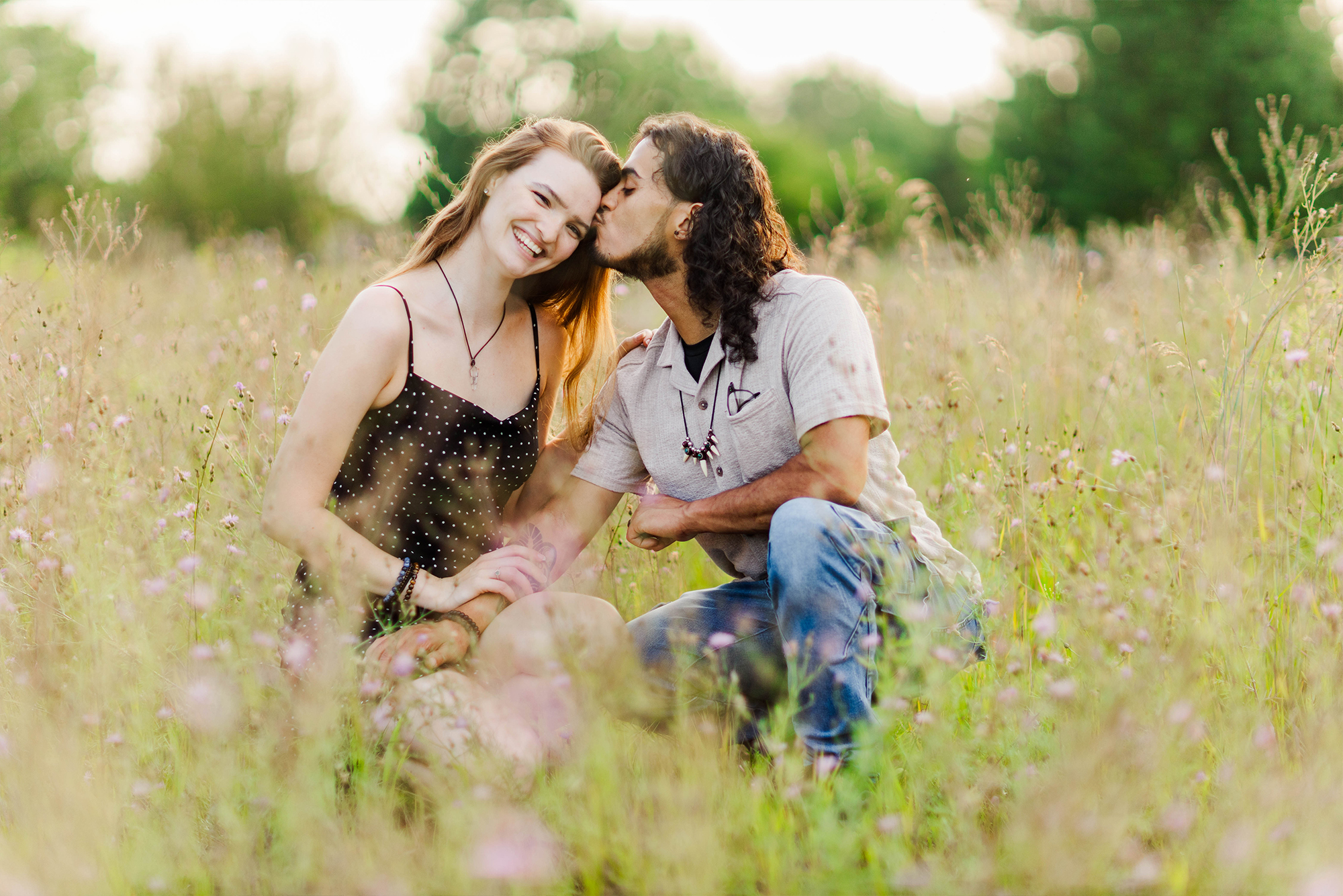 boyfriend and girlfriend sitting in tall grass sharing a kiss during sunset