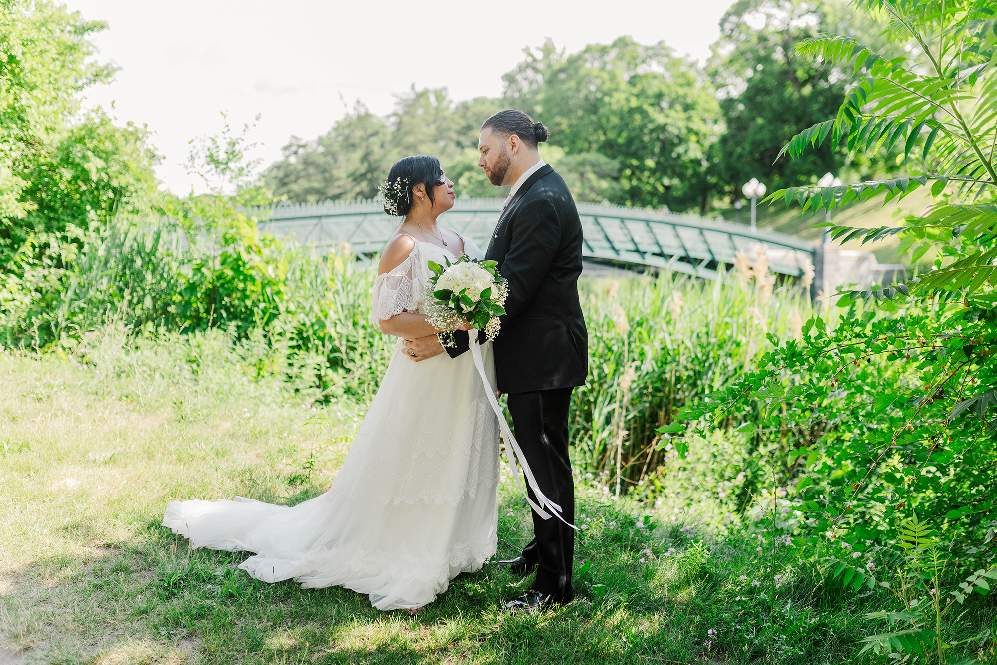 bride and groom posing in park with bridal bouquet with bridge backdrop