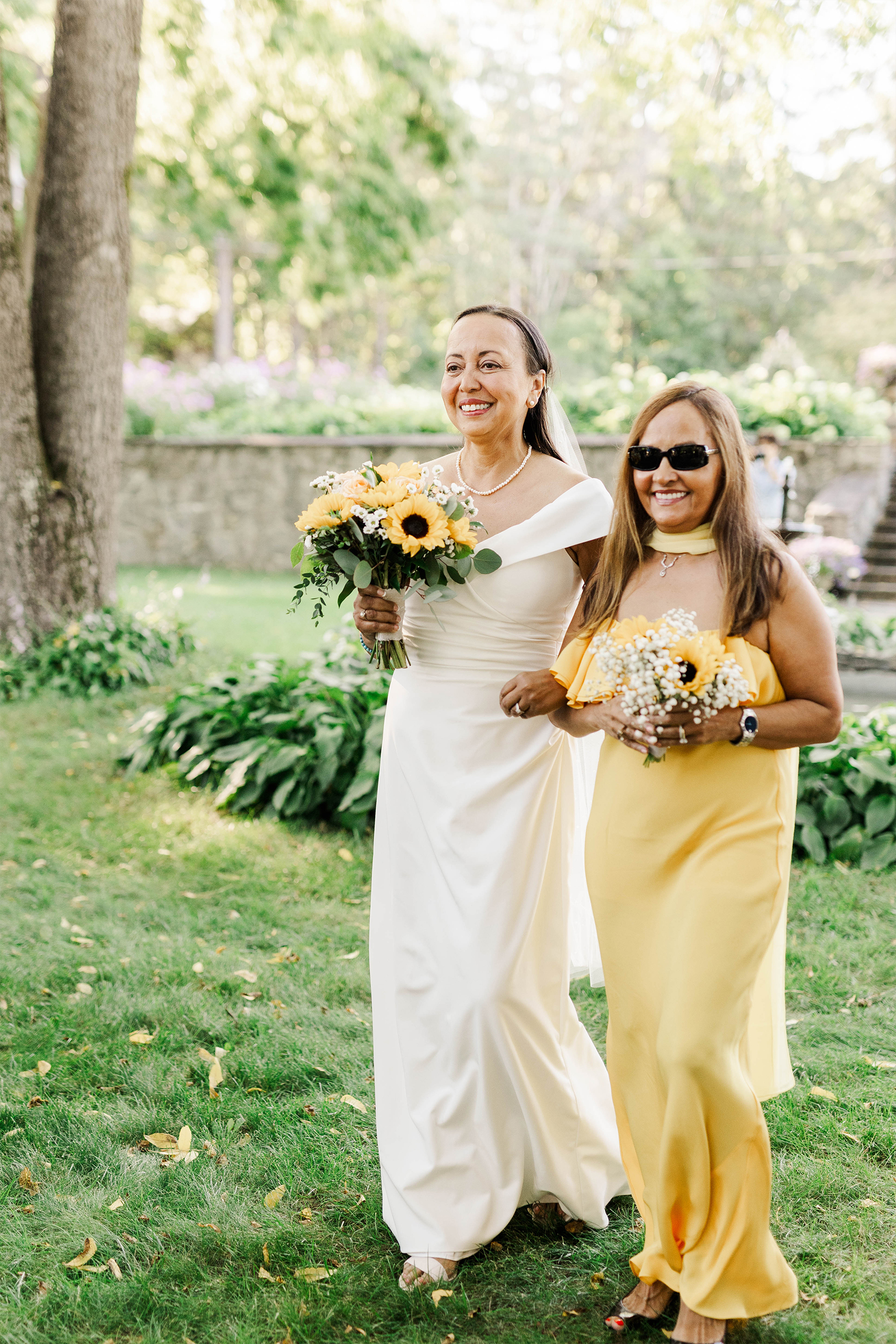 bride walking down aisle with. maid of honor both holding bouquet of sunflowers 