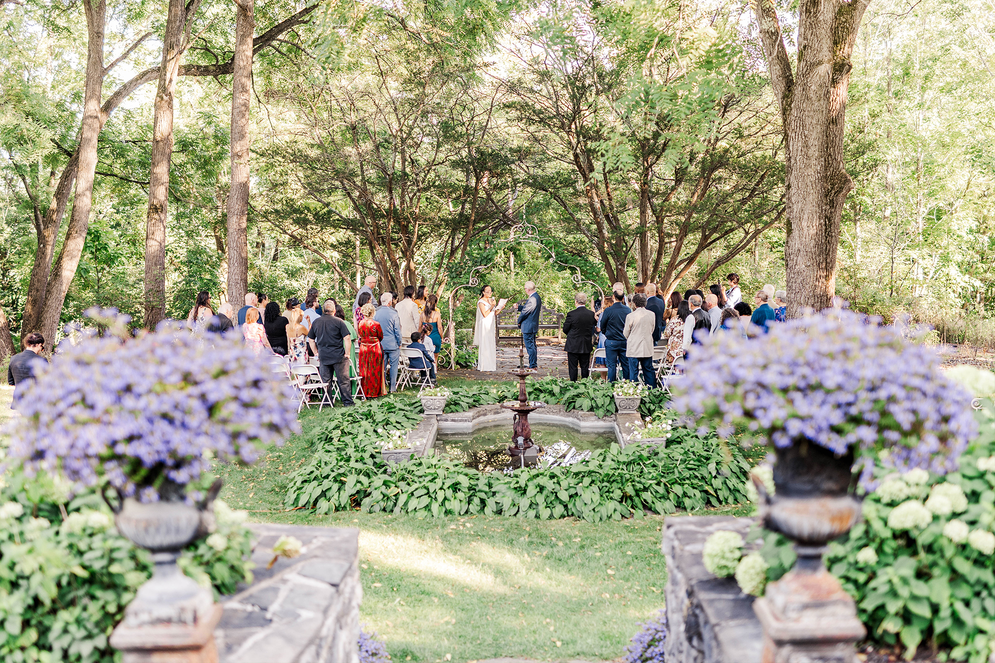 bride making a speech to groom underneath wedding arch surrounded by guests ceremony space and flowers