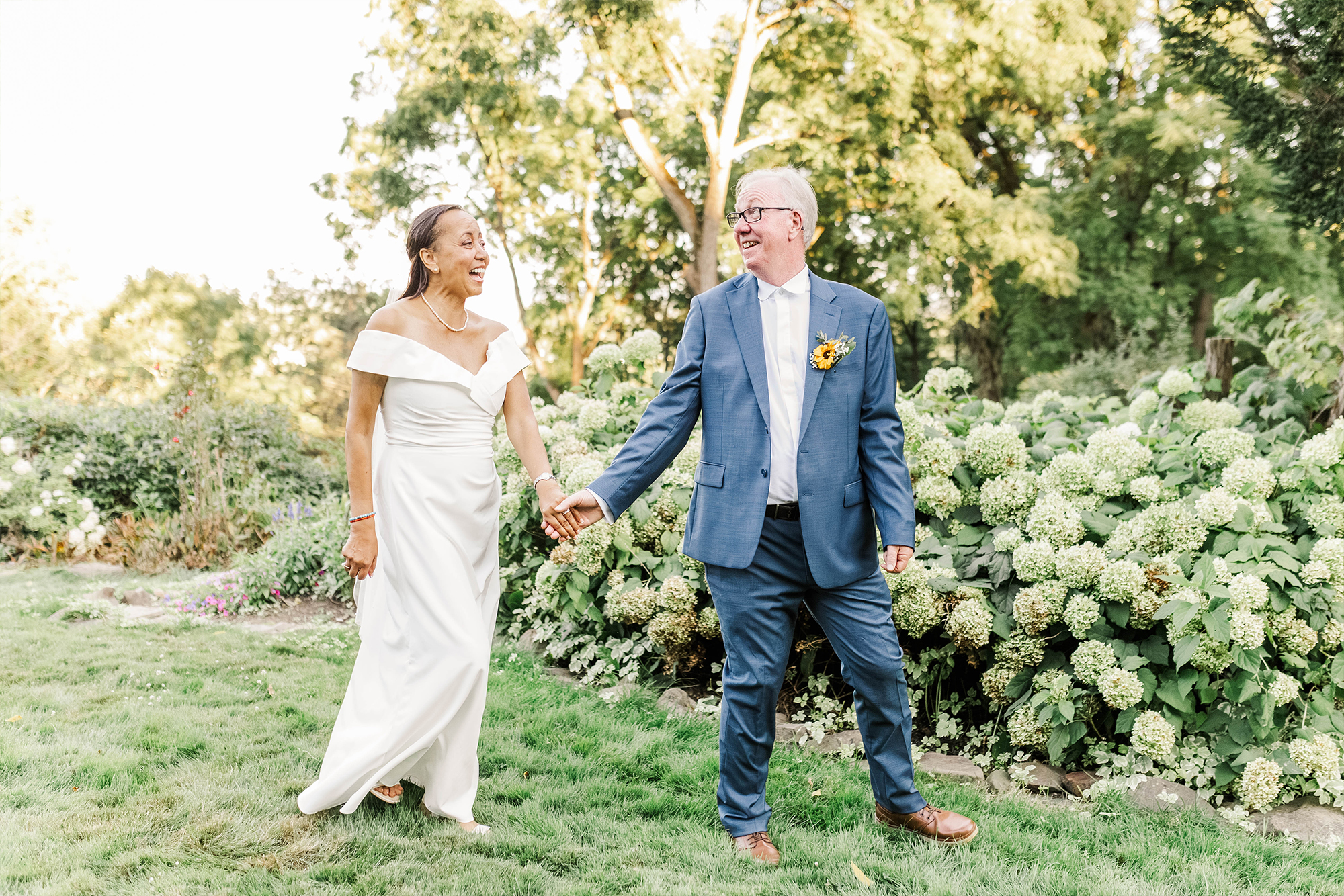 groom walking ahead of bride while holding her hand as she follows on grass with trees and greenery
